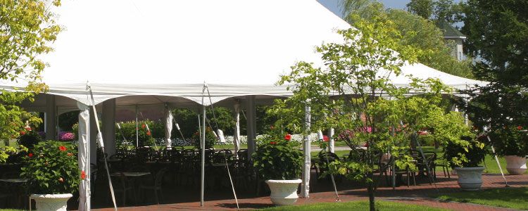 A white party tent set up with tables and chairs in a garden by Pinnacle Rental Centers